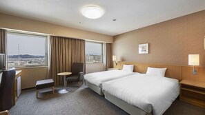 Habitación de hotel con dos camas, un escritorio, una silla, una mesita y vistas a la ciudad.