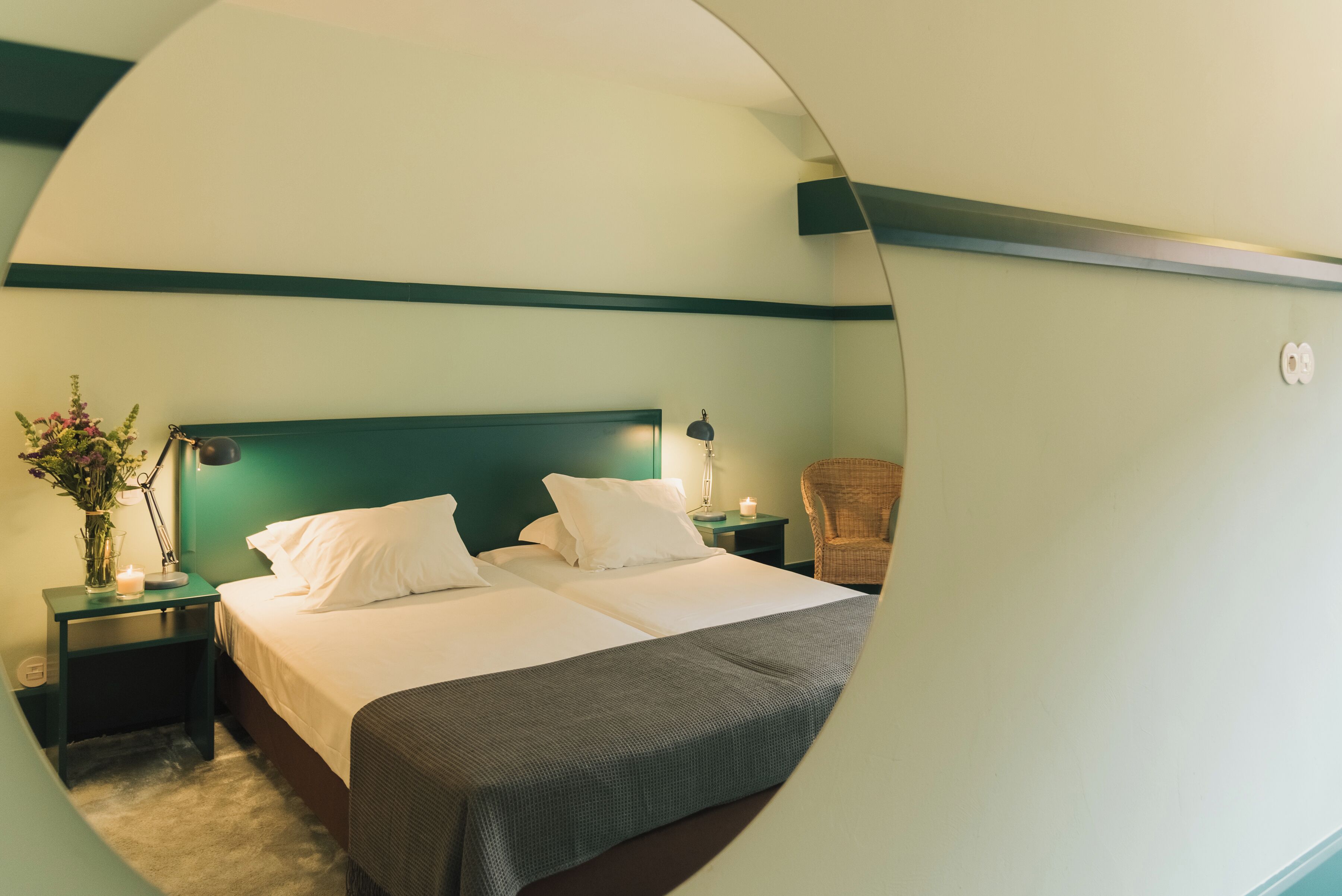 En panoramavy över ett hotellrum med en säng, ett nattduksbord, en lampa, en stol och en vas med blommor.