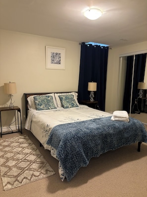 Un dormitorio con una cama, dos mesitas de noche, una lámpara, una alfombra, un cuadro enmarcado y una ventana con cortinas.