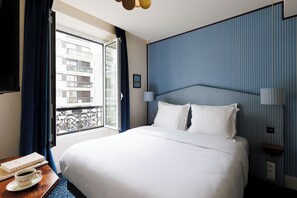 Une chambre d’hôtel avec un lit, une fenêtre avec des rideaux, une table de chevet avec une tasse et un livre, et un mur orné d’un motif à rayures.