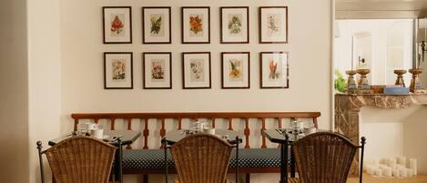 A dining area with a bench and chairs, framed botanical prints on the wall, and a fireplace with candles.