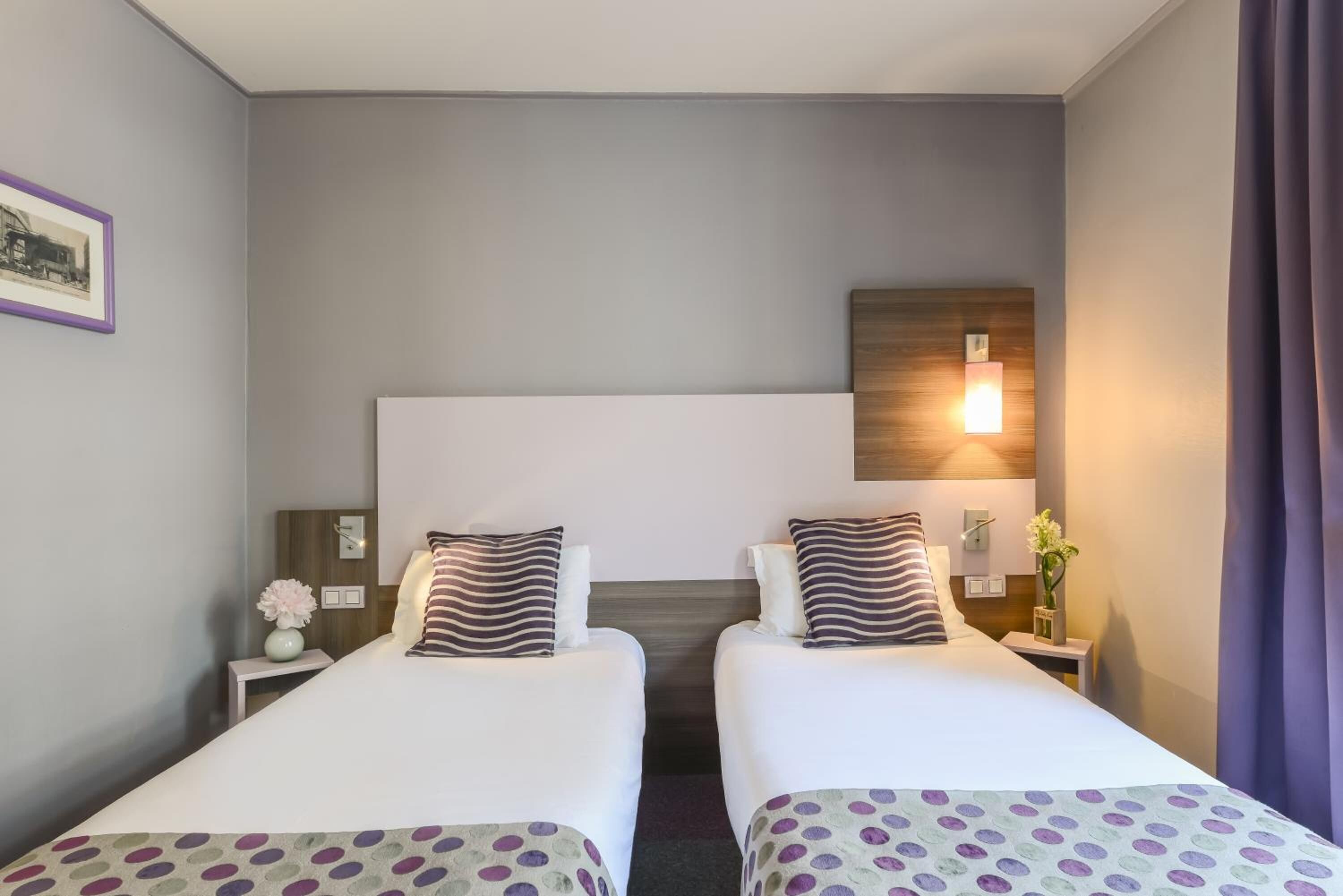 A hotel room with two beds, a wooden headboard, a nightstand with a vase, and a wall-mounted light.