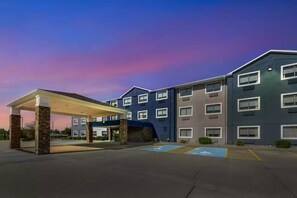 A multi-story hotel with a covered entrance, parking spaces, and a clear sky.