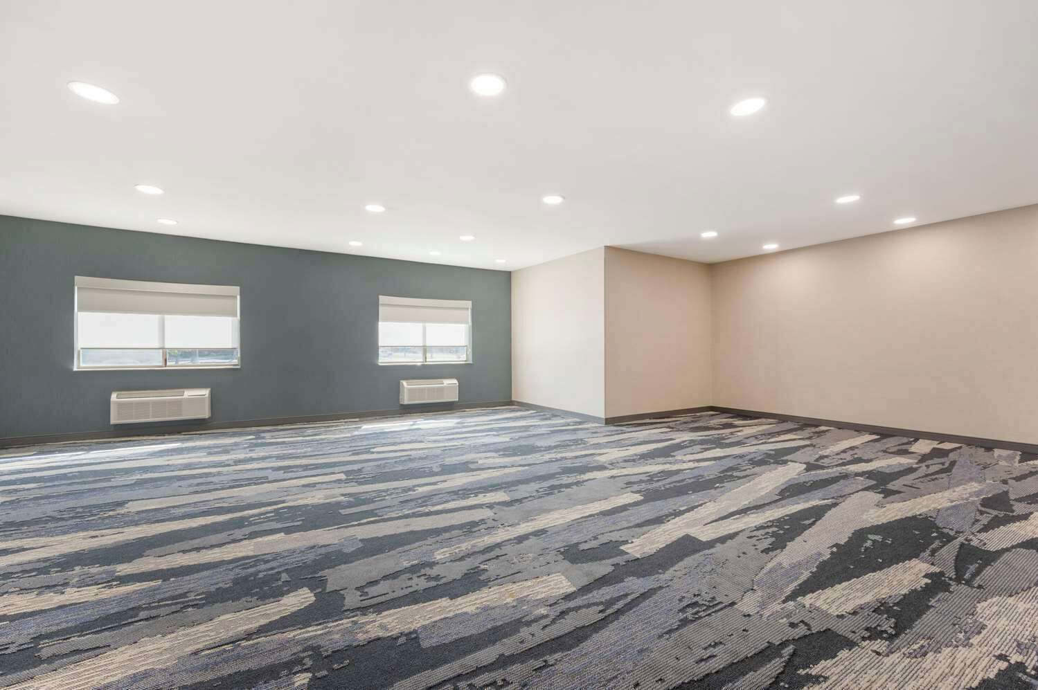 A large, empty room with a patterned carpet, two windows with blinds, and a ceiling with recessed lighting.
