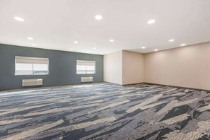 Una habitación amplia y vacía con alfombra estampada, dos ventanas con persianas y un techo con iluminación empotrada.