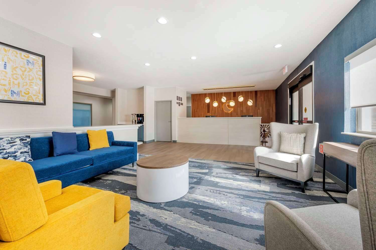 A modern living room with a blue sofa, a yellow armchair, a white sofa, and a coffee table. There is a kitchen area with wooden cabinets and pendant lighting.