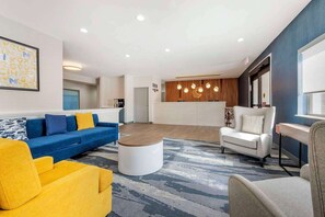A modern living room with a blue sofa, a yellow armchair, a white sofa, and a coffee table. There is a kitchen area with wooden cabinets and pendant lighting. - Comfort Inn Kendallville East, IN (Kendallville)