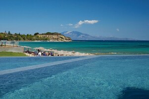 Zona de piscina con agua azul cristalina, playa con sombrillas y una montaña al fondo.
