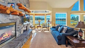 A living room with a stone fireplace, a large window with a view of a lake, a blue sofa, and a flat-screen TV. - Big Bear Lakefront Luxury Home w/ Spa & Views (Big Bear Lake)
