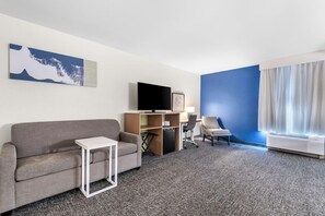 Ein Hotelzimmer mit grauem Sofa, einem kleinen weißen Tisch, einem Fernseher auf einem Holzständer, einem Schreibtisch mit Stuhl und einem großen Fenster mit Vorhängen.