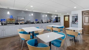 Un espace repas moderne avec des tables blanches et des chaises bleues, un buffet proposant différents plats, et un mur décoratif à motifs.