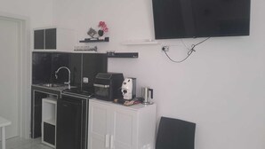 Petita cuina compacta amb fornet, rentavaixelles i cafetera, un televisor de pantalla plana fixat a la paret i una cadira.