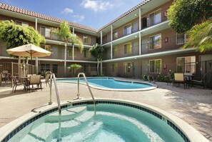 Una piscina con una escalera metálica, rodeada de mobiliario exterior y un edificio de varios pisos con balcones.