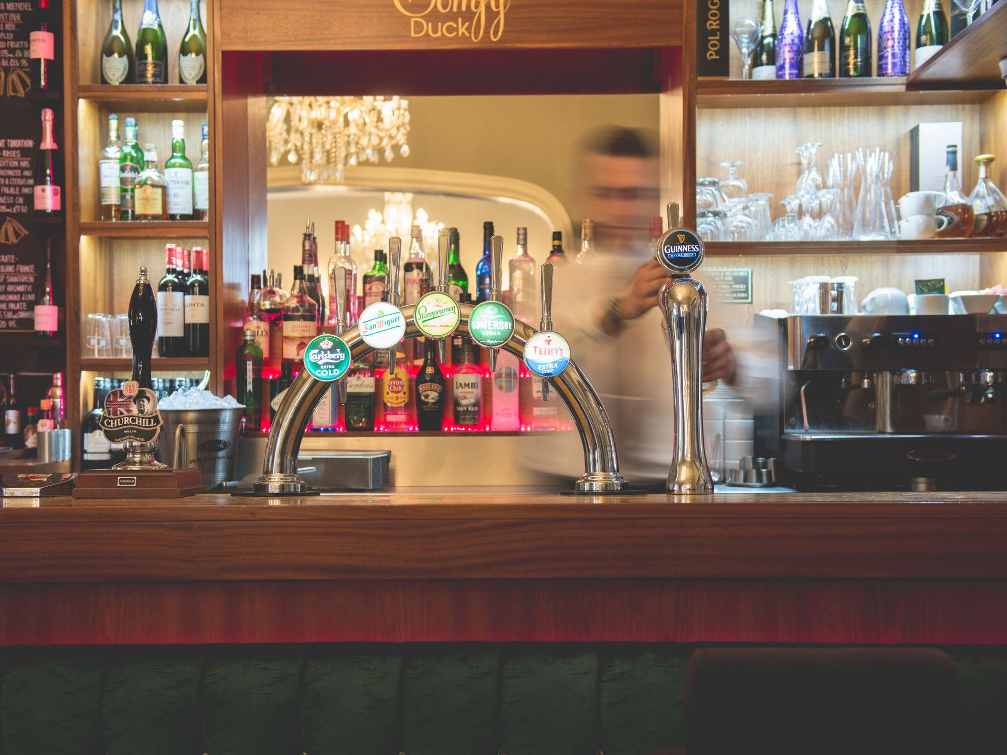 A bar with multiple taps, a variety of bottles, and a blurred person in the background.