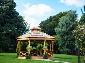 Um gazebo de madeira com telhado de cerâmica, cercado por um gramado bem cuidado e árvores densas.