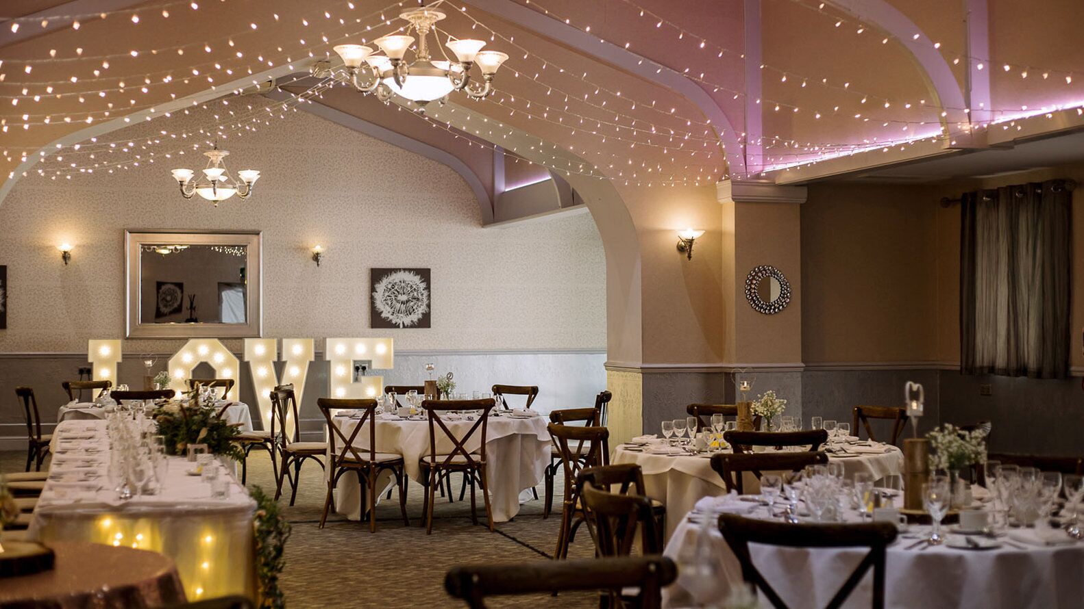 A banquet hall with multiple round tables, chandeliers, and string lights.