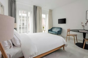 A hotel room with a bed, a desk, a chair, a TV, and large windows with curtains. - Sonder Santa Ana (Madrid)