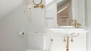 A modern bathroom with a skylight, a toilet, a sink with gold fixtures, and a large mirror. - Sonder Santa Ana (Madrid)