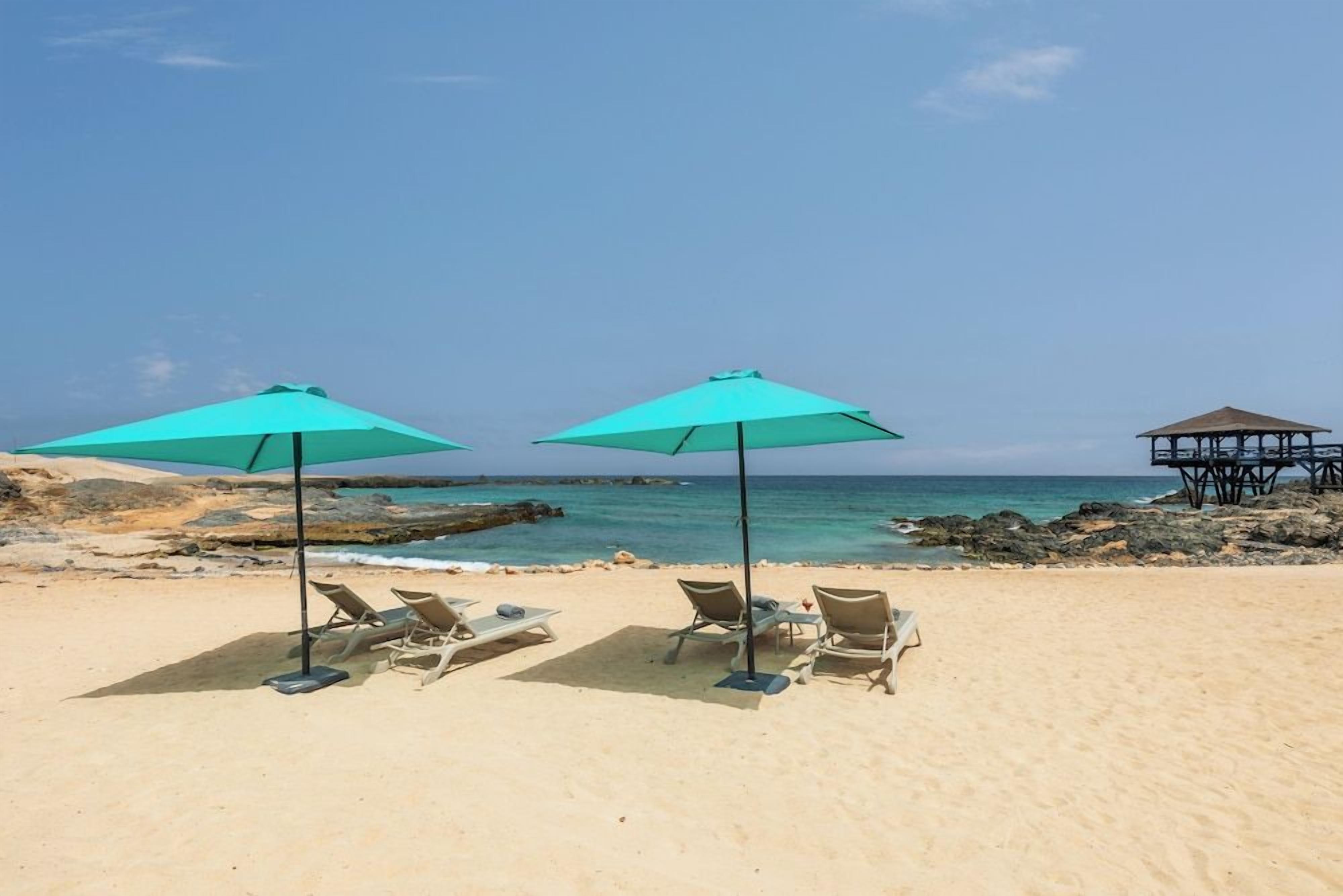 A beach with two turquoise umbrellas and lounge chairs, a rocky shoreline, and a wooden structure in the background.