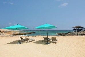 A beach with two turquoise umbrellas and lounge chairs, a rocky shoreline, and a wooden structure in the background. - Ahg Marine Club Beach Resort (Boa Vista)