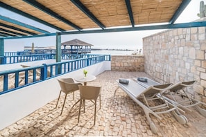 Un patio exterior con una mesa y sillas, una pequeña mesa redonda con una planta, y una zona de estar con un banco.