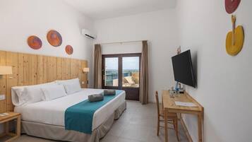 A hotel room with a bed, a desk, a TV, and a view of the sea.
