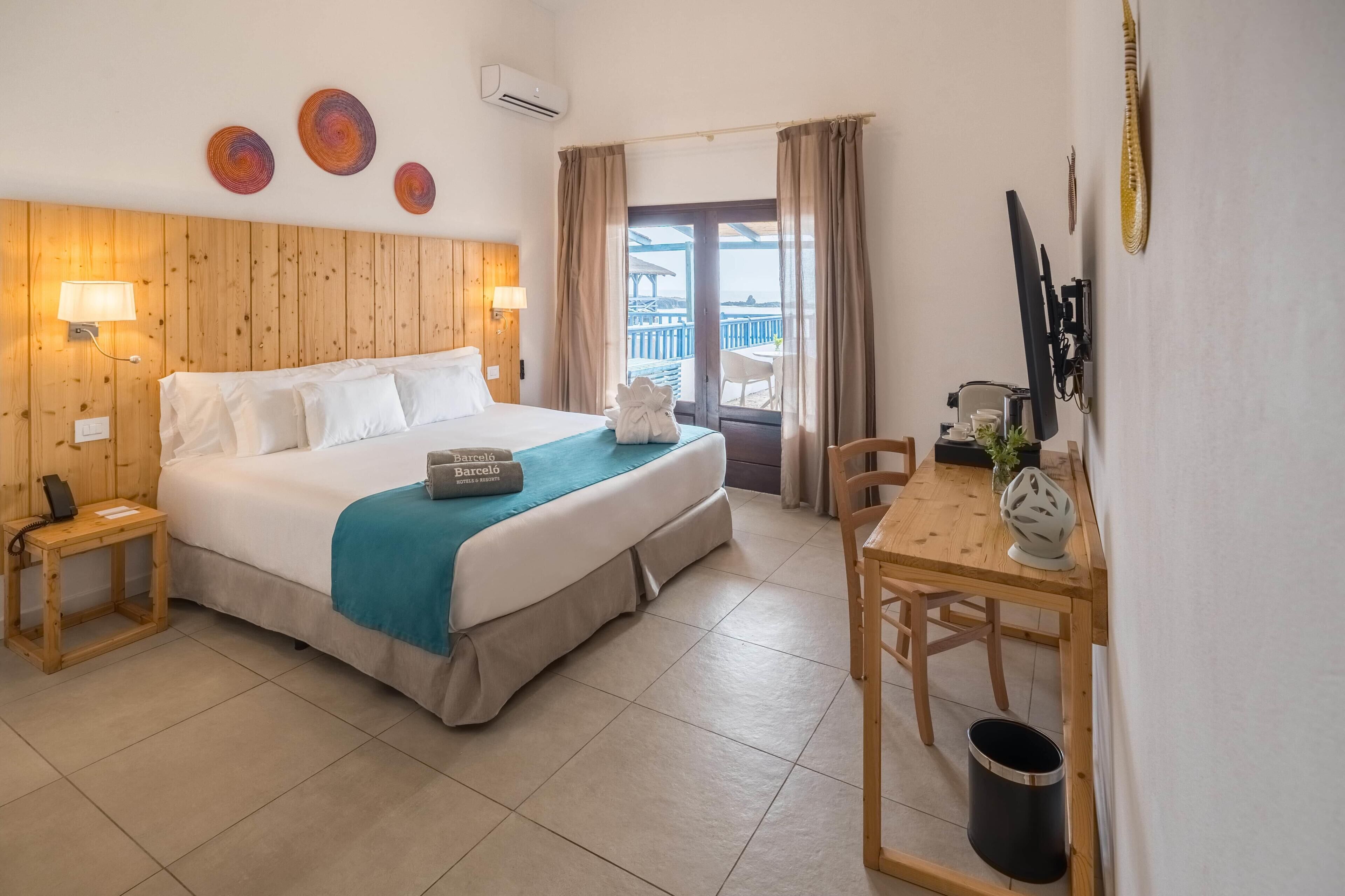 A hotel room with a bed, bedside tables, a desk with a chair, a window with curtains, and a view of the sea.