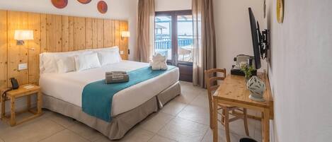 Habitación de hotel con cama, mesitas de noche, un escritorio con silla, ventana con cortinas y vista al mar.