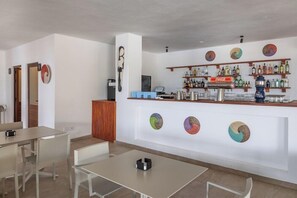 A bar area with shelves of bottles and a Coca-Cola sign, a seating area with tables and chairs, and decorative wall art. - Ahg Marine Club Beach Resort (Boa Vista)