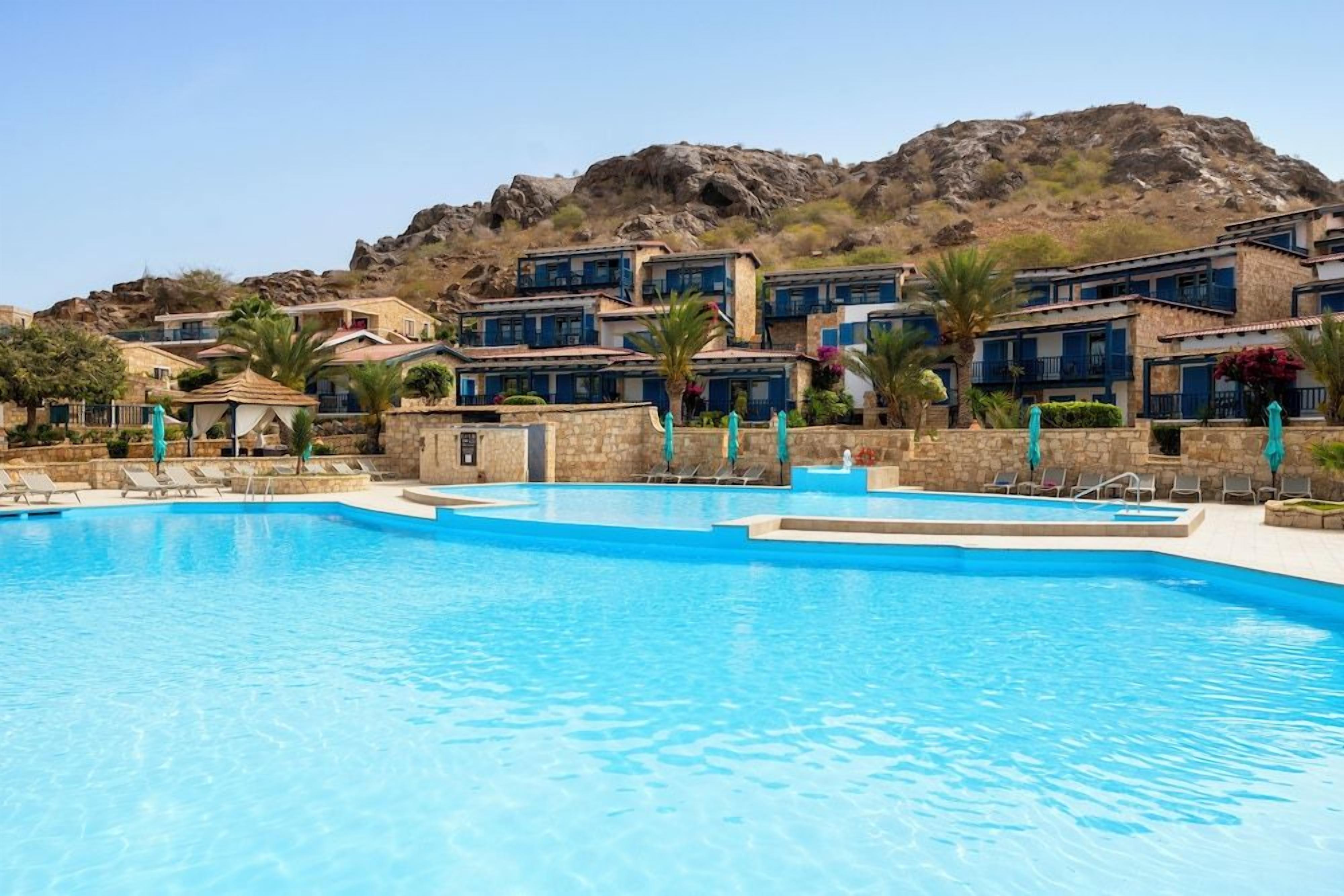 Una piscina con sillas de descanso, montañas al fondo y edificios con detalles azules.