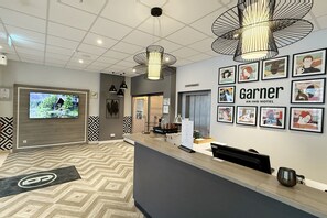 A hotel reception area with a modern design, including a reception desk, framed artwork on the wall, and a television screen displaying a scenic view. - Yggotel Ravn (Berlin)