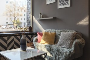A cozy room with a sofa, a vase with flowers, a coffee table, and a window with a view of buildings and trees. - Yggotel Ravn (Berlin)