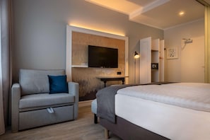 A modern hotel room with a bed, a sofa, a TV mounted on the wall, a desk, and a door with a mirror. - Yggotel Ravn (Berlin)