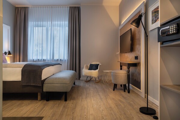 Ein Hotelzimmer mit einem Bett, einem Sessel, einem kleinen Tisch, einer Stehlampe und einem an der Wand befestigten Fernseher.