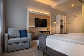 A modern hotel room with a bed, a sofa, a TV mounted on the wall, a bedside table, and a door with a mirror. - Yggotel Ravn (Berlin)