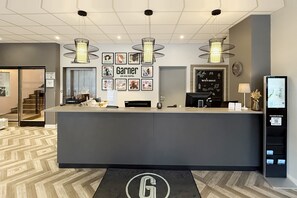 Un comptoir d'accueil d'hôtel au design moderne, avec des œuvres d'art encadrées accrochées au mur, et un kiosque d'information numérique situé à droite.