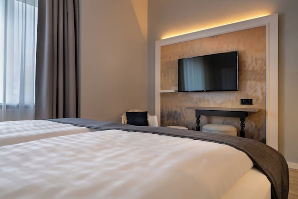 A hotel room with two beds, a flat-screen TV mounted on the wall, a small table, and a window with curtains. - Yggotel Ravn (Berlin)