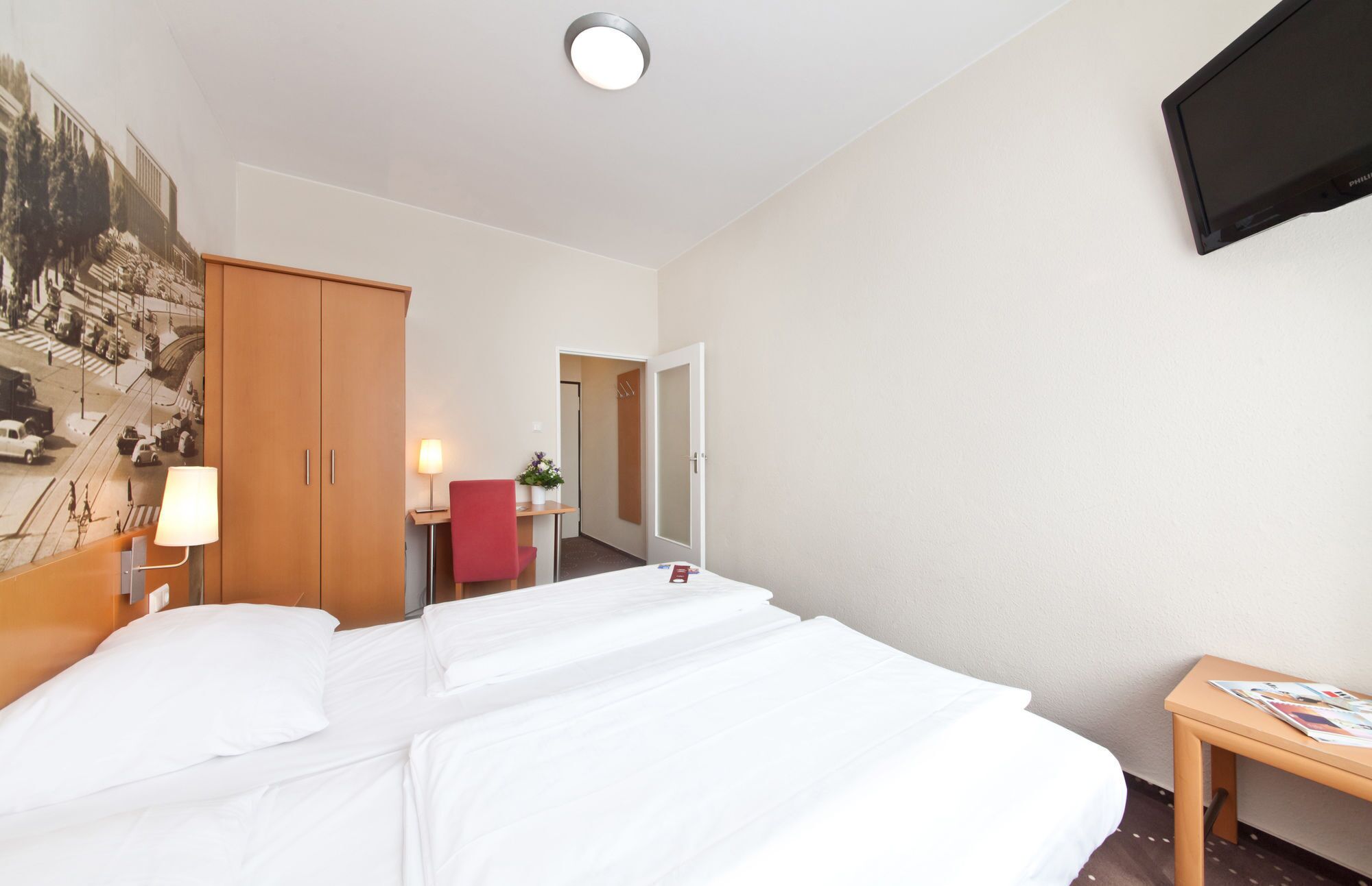 Quarto de hotel com cama, escrivaninha, cadeira, abajur, televisão e porta.