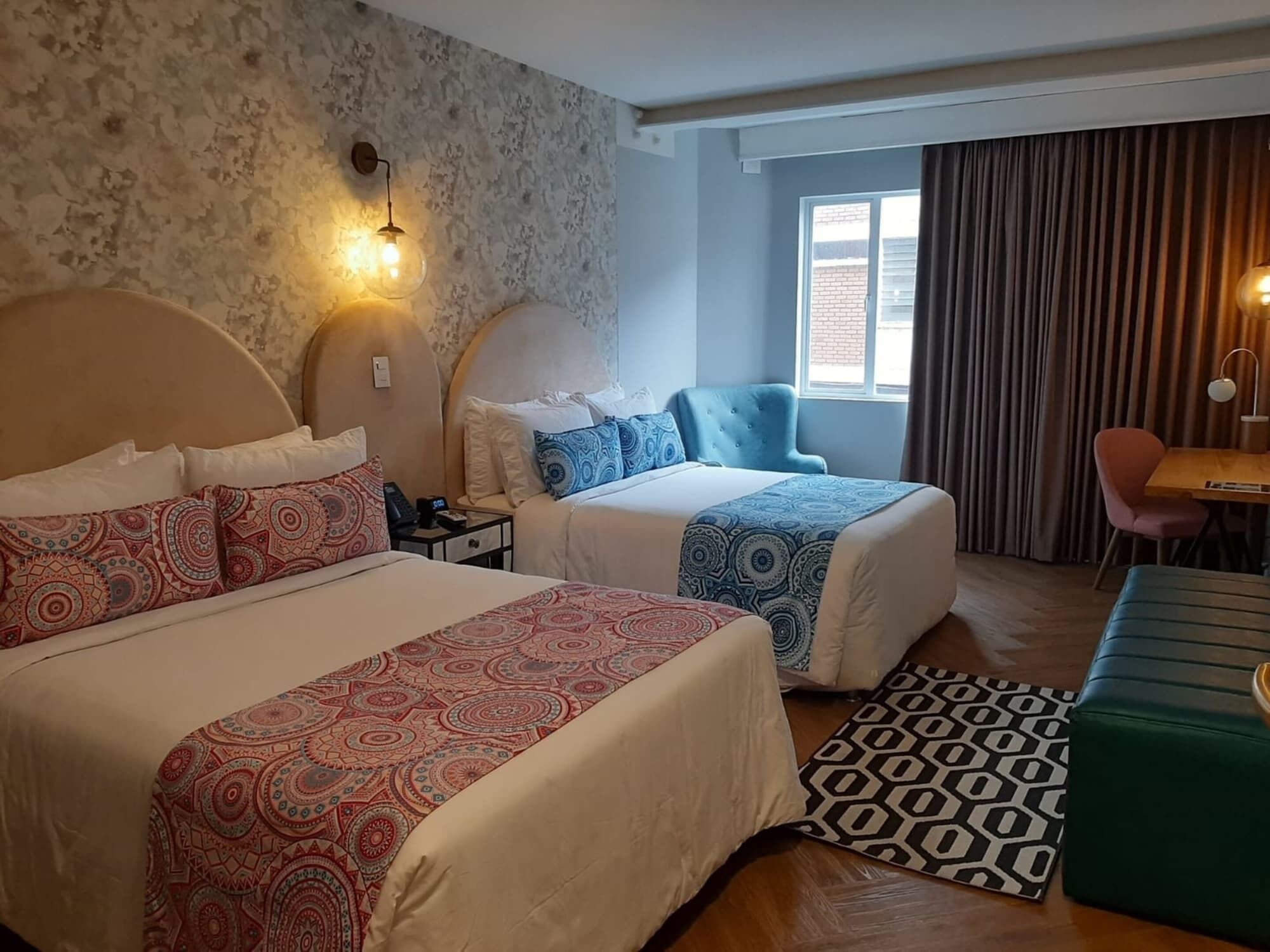 Une chambre d’hôtel avec deux lits, un bureau, une chaise, un tapis à motifs et une fenêtre avec des rideaux.