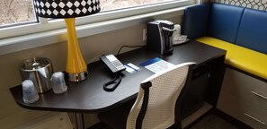 Ein kleiner Schreibtisch mit Telefon, Kaffeemaschine, Lampe und einem Fenster mit Blick ins Freie.