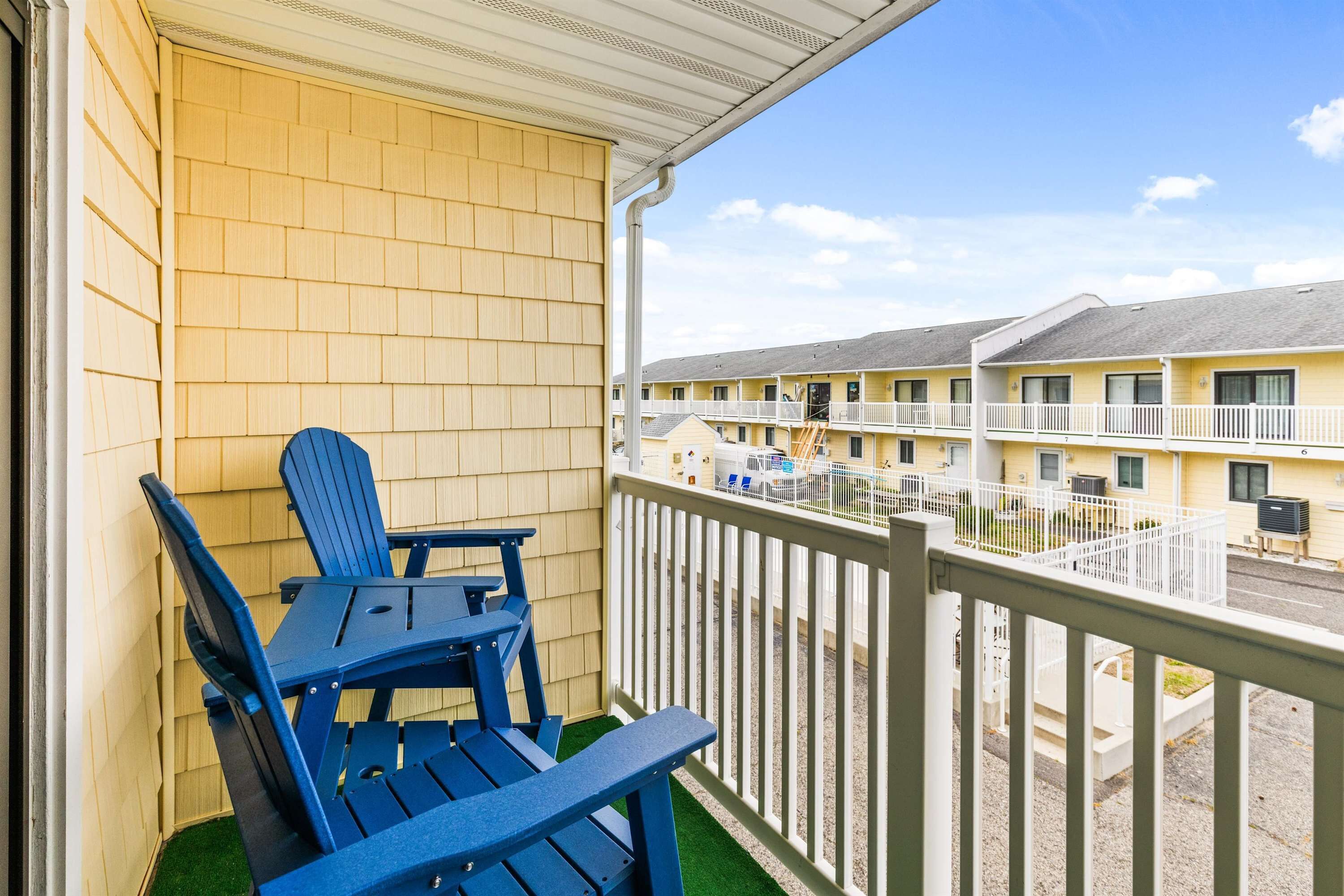 Un balcon agrémenté de chaises Adirondack bleues, un bâtiment jaune avec une balustrade blanche, et une vue sur un motel doté de nombreux balcons.