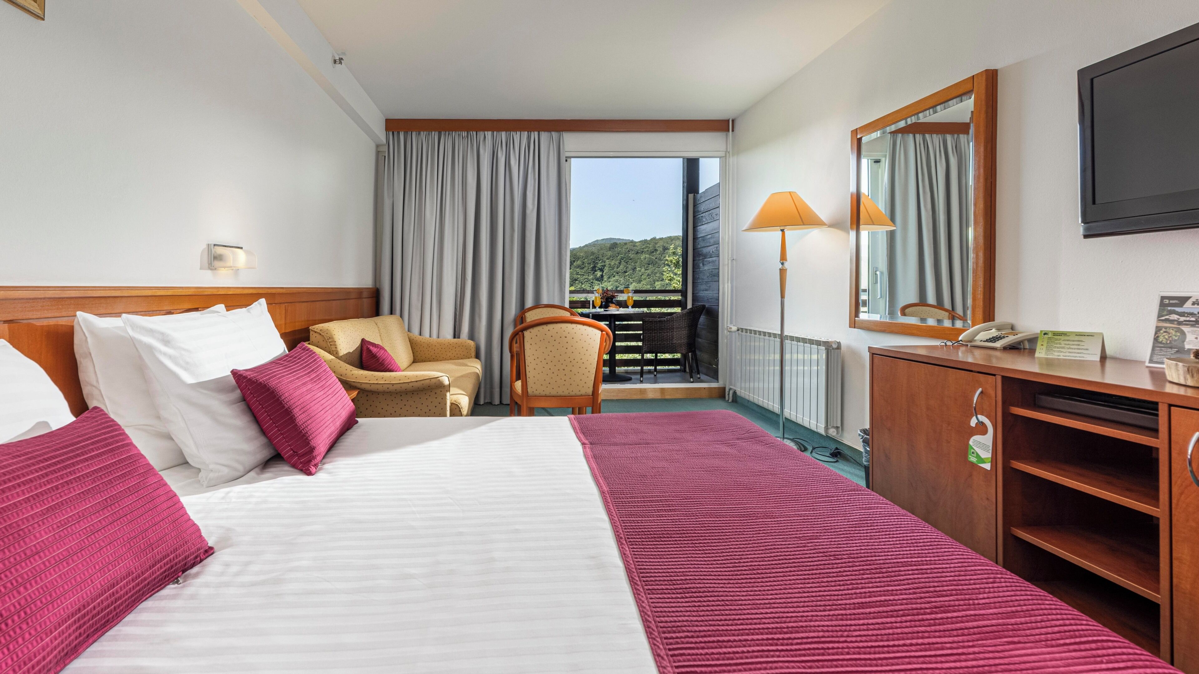 A hotel room with a bed, a desk, a chair, a balcony with a view, and a mirror.