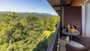 Ein Balkon mit Tisch, einem Korbstuhl und Blick auf eine bewaldete Landschaft.