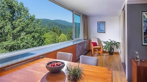 Ein Zimmer mit großem Fenster und Blick auf einen bewaldeten Berg, einem Holzesstisch mit Obstschale und einem Stuhl.