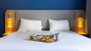 Una cama bien hecha con sábanas blancas, dos almohadas, una bandeja con desayuno que incluye un croissant, fruta y una taza, y lámparas de noche.