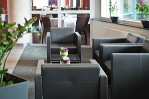Eine moderne Lounge mit Ledersitzgelegenheiten, einem kleinen Tisch mit Blumenarrangement und Blick auf ein Restaurant im Hintergrund.