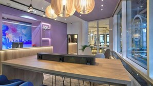 A modern reception area with a bar, a large screen displaying a cityscape, and a seating area with blue chairs. - Intercityhotel Kassel (Kassel)