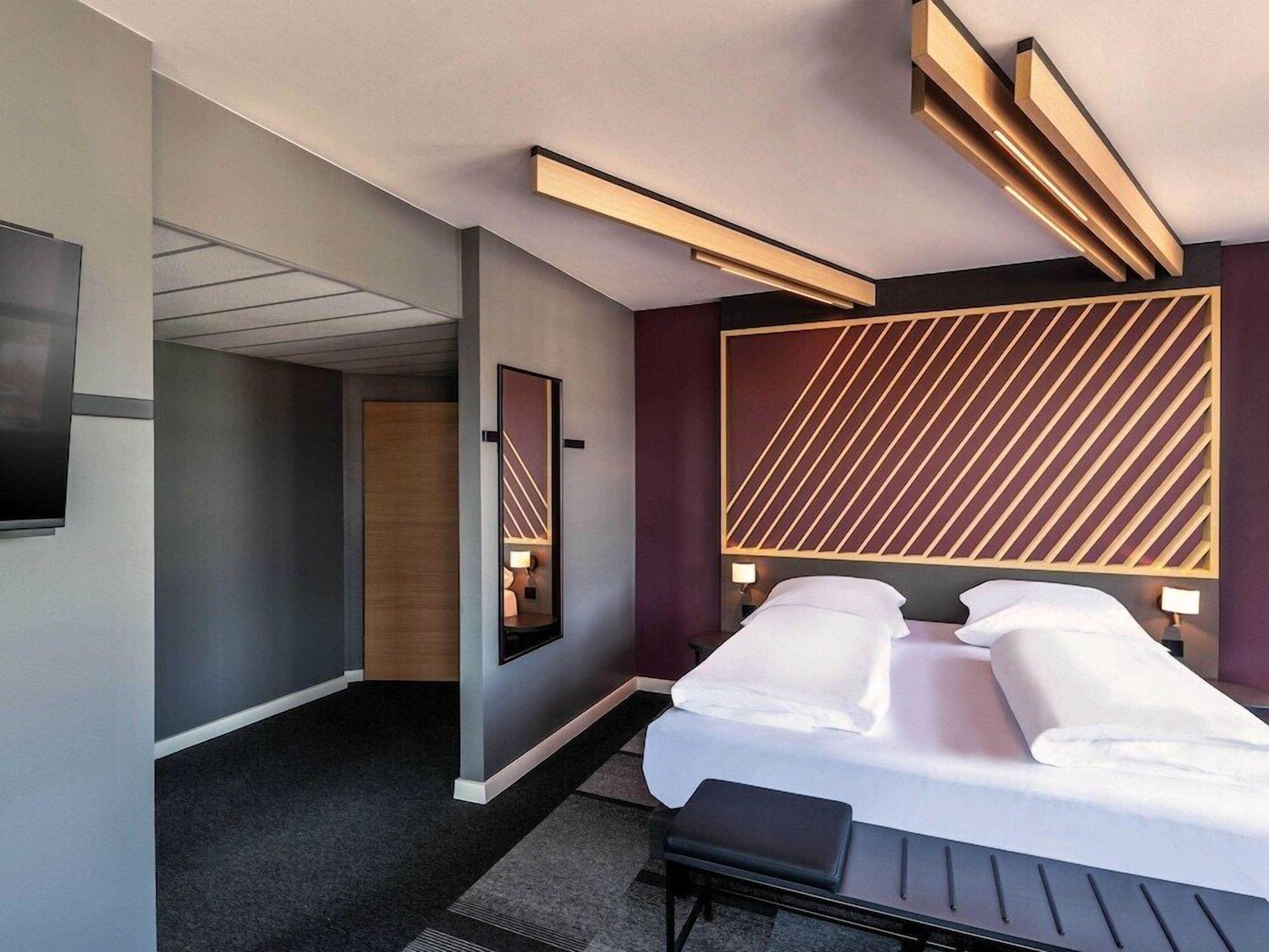 A modern hotel room with a large bed, a flat-screen TV, and a unique geometric wall design.