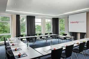 A conference room with a large screen displaying 'Intercity Hotel,' multiple tables with chairs, and large windows with curtains. - Intercityhotel Kassel (Kassel)
