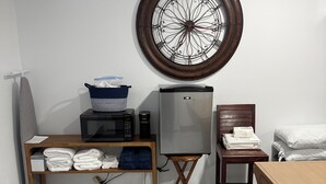 A room with a bed, a table with towels, a microwave, a refrigerator, a chair, and a wall clock. - Cozy 1-bedroom cottage in serene Ravenel with AC (Ravenel)